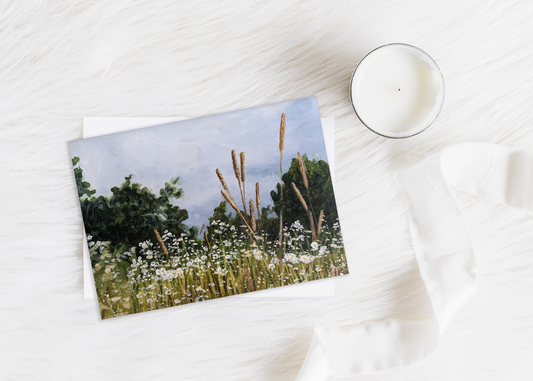 A greeting card with an acrylic painting of a field of tall grass under a stormy sky, presented on a white surface with a candle and a white furry surface in the background.