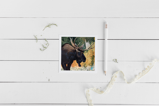 A greeting card featuring an acrylic painting of a bull moose in autumn foliage, accompanied by a brown kraft envelope and a pencil, displayed on a white wooden background with lace.