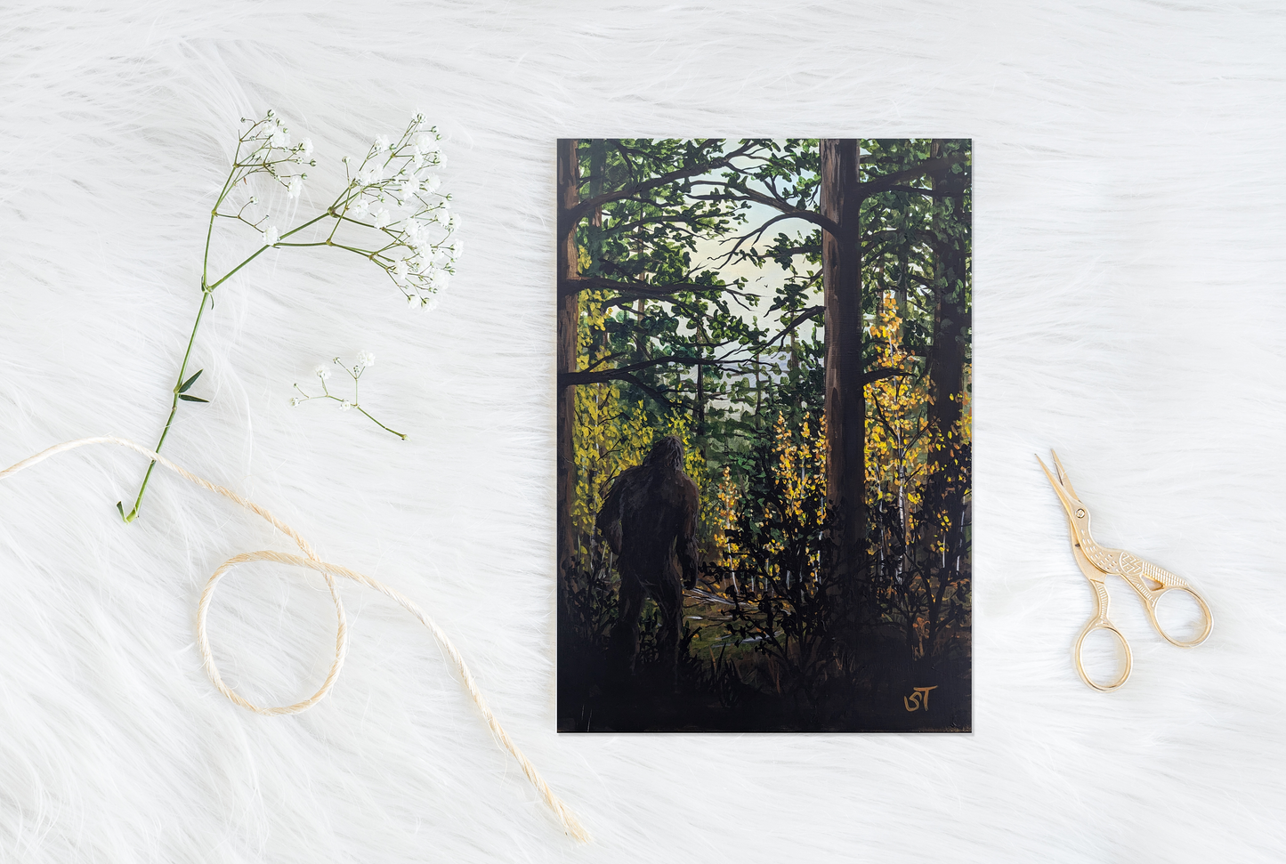 Sasquatch illustration walking through a forest, shown as a greeting card styled on a white surface with scissors, twine, and delicate dried flowers.