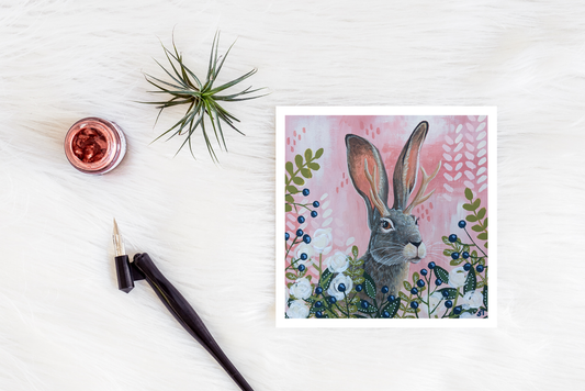 Jackalope illustration with antlers among flowers and berries on a pink background, shown as a greeting card styled on a white surface with art tools and greenery.