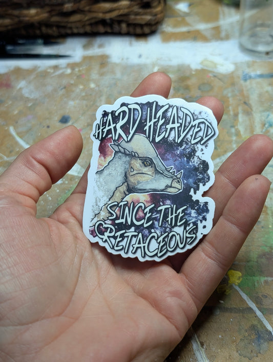 Sticker with a pachycephalosaurus design held in a hand on a textured surface.