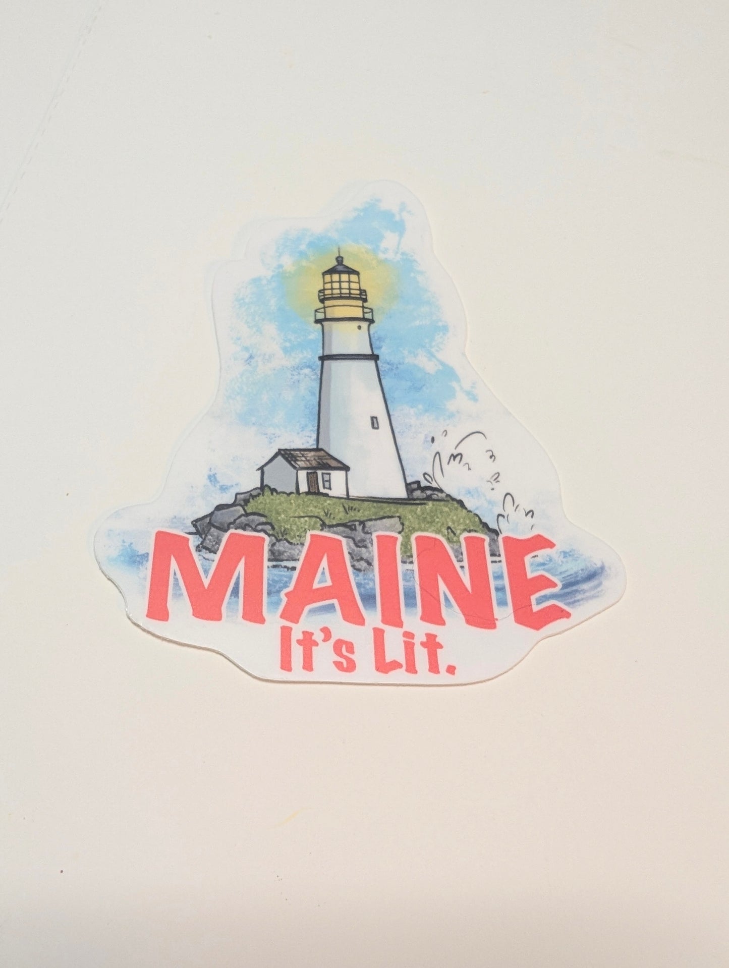 βMaine itβs litβ lighthouse vinyl sticker shown in a flat lay on a white background.