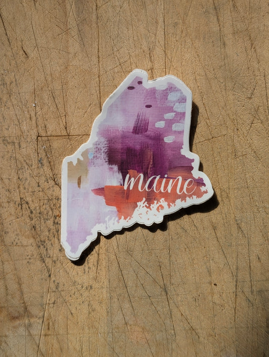 Abstract pink and purple Maine-shaped vinyl sticker with “maine” text, shown in a flat lay on wood.