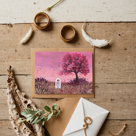Greeting card with a pink tree and ghost illustration on a wooden surface with decorative items.