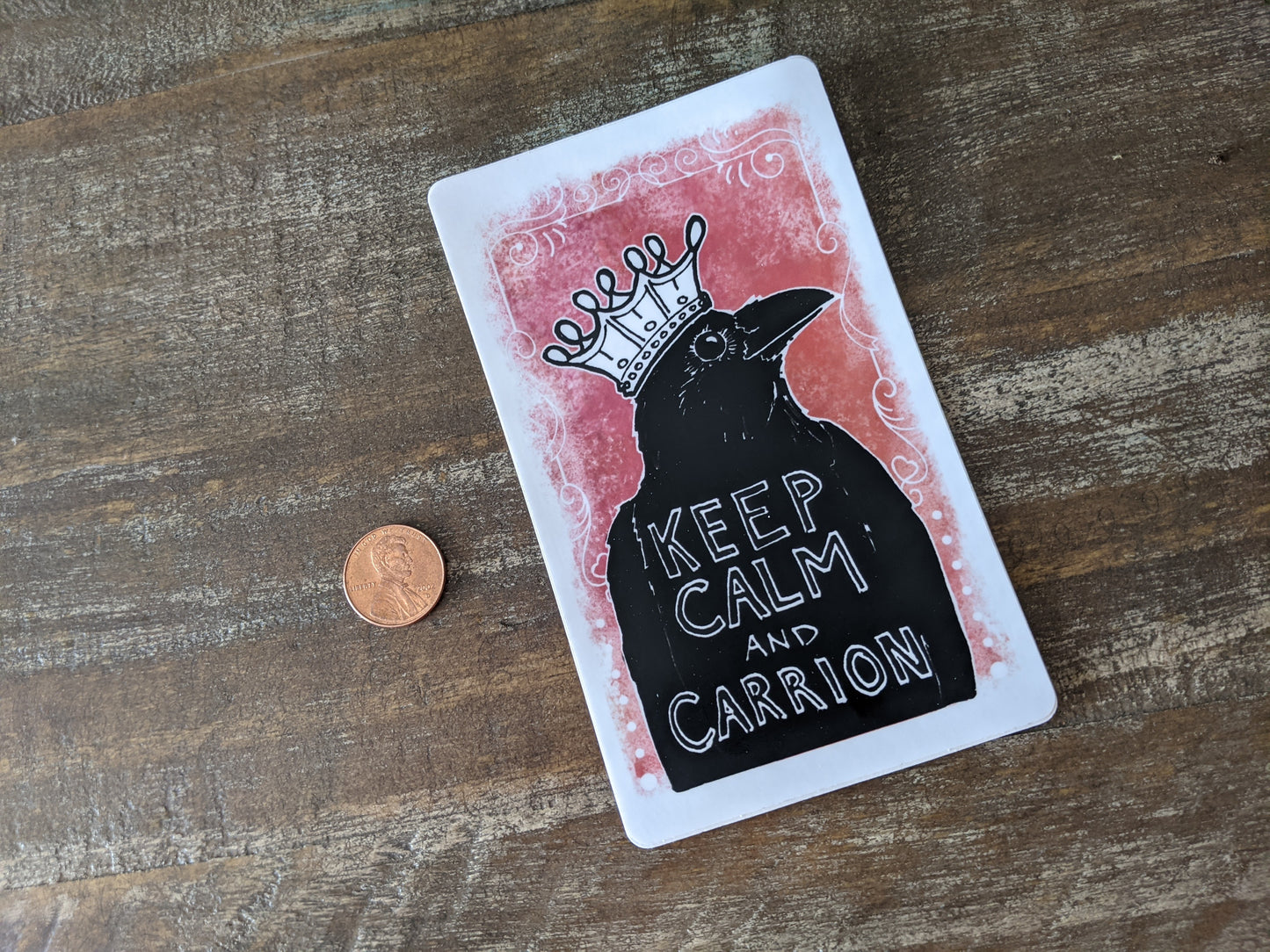 Raven vinyl sticker with a crown and the text “Keep calm and carrion,” shown next to a penny for size reference on a wooden surface.