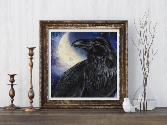 Framed art print of a black raven before a glowing full moon, styled on a wooden surface with brass candlesticks and a glass vase of bare branches.