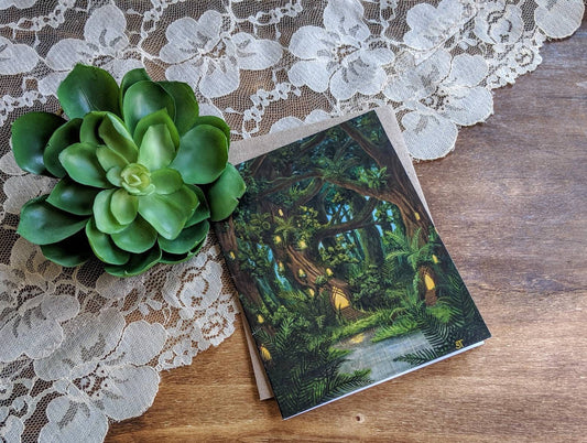 Greeting card showing a lush fantasy forest with glowing fairy house lights among towering trees, styled on a wooden surface with lace fabric and a green succulent.