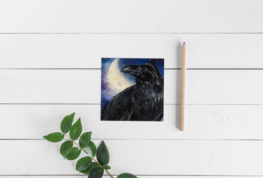 Square greeting card showing a painting of a black raven in profile against a moonlit night sky, photographed on a white wood surface with a pencil and green leafy branch.