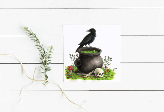 Square greeting card featuring a black raven perched on a bubbling cauldron, with a skull, red mushroom, and greenery at the base, styled on a white wood surface with a sprig of greenery and twine.