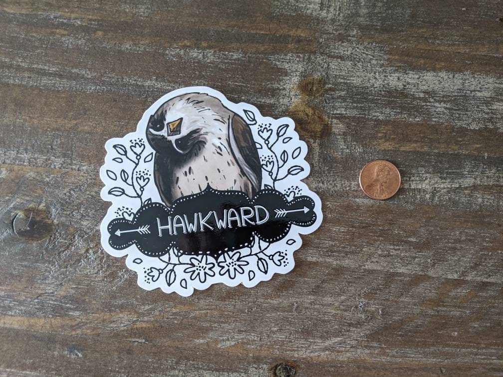 A sticker with an illustration of an awkward hawk and the word 'HAWKWARD' written on it, placed on a wooden surface, next to a penny for scale.