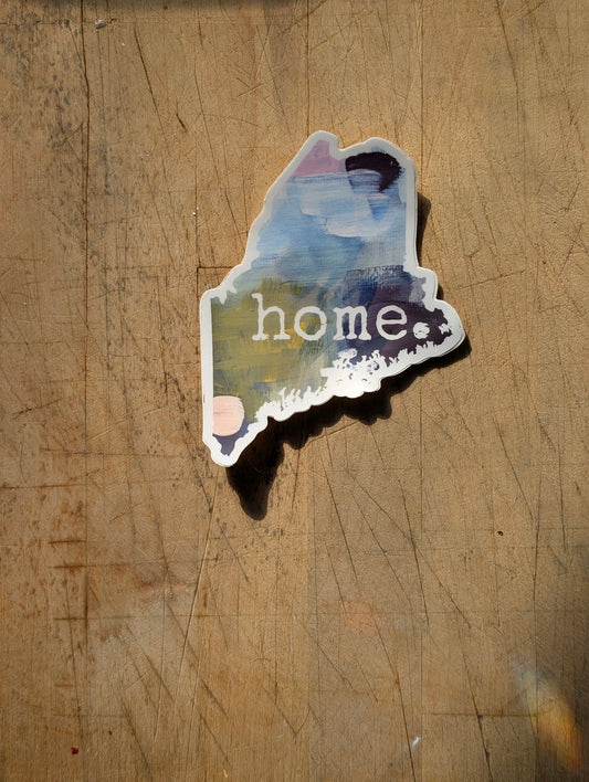 Abstract Maine-shaped “home” vinyl sticker with blue, green & pink acrylic textures, shown in a flat lay on a wooden surface.