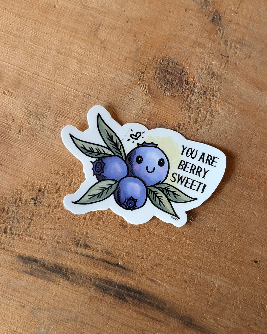 A vinyl sticker with a illustration of a smiling blueberry with leaves and the phrase 'YOU ARE BERRY SWEET' written alongside.