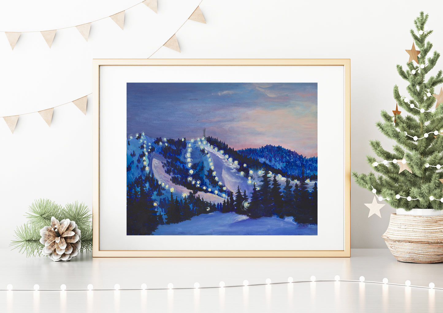 A framed art print depicting a night scene of Bogus Basin with snow-covered mountains and ski slopes, displayed on a white wall with decorative items around it.