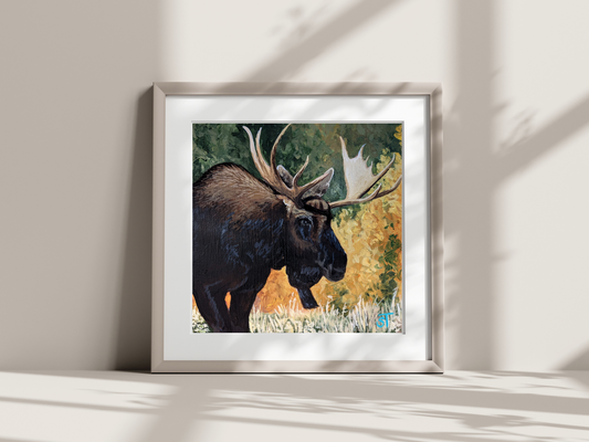 A framed art print of a moose with a forest backdrop, displayed on a wall in a room with natural lighting.