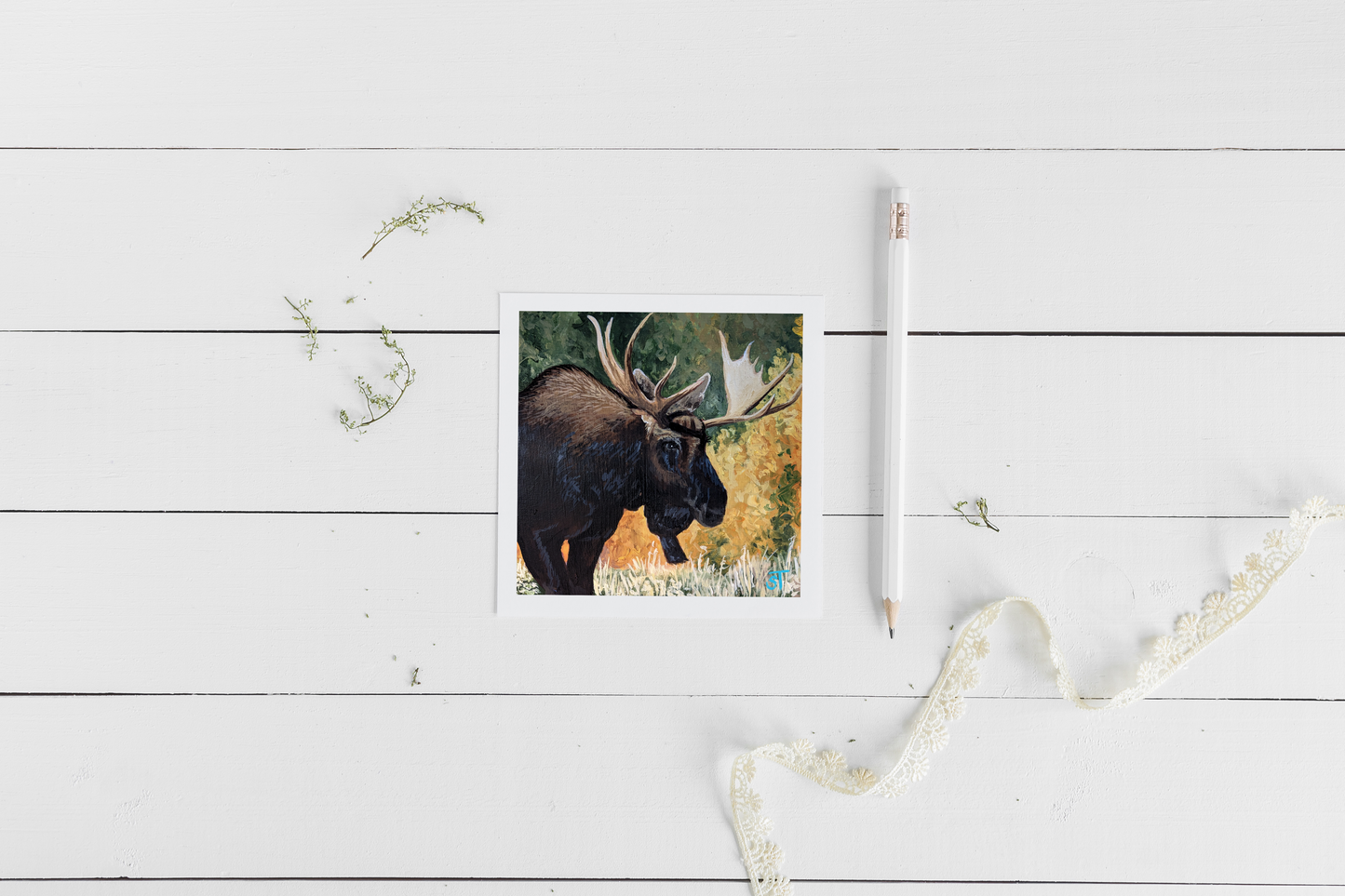 A greeting card featuring an acrylic painting of a bull moose in autumn foliage, accompanied by a brown kraft envelope and a pencil, displayed on a white wooden background with lace.