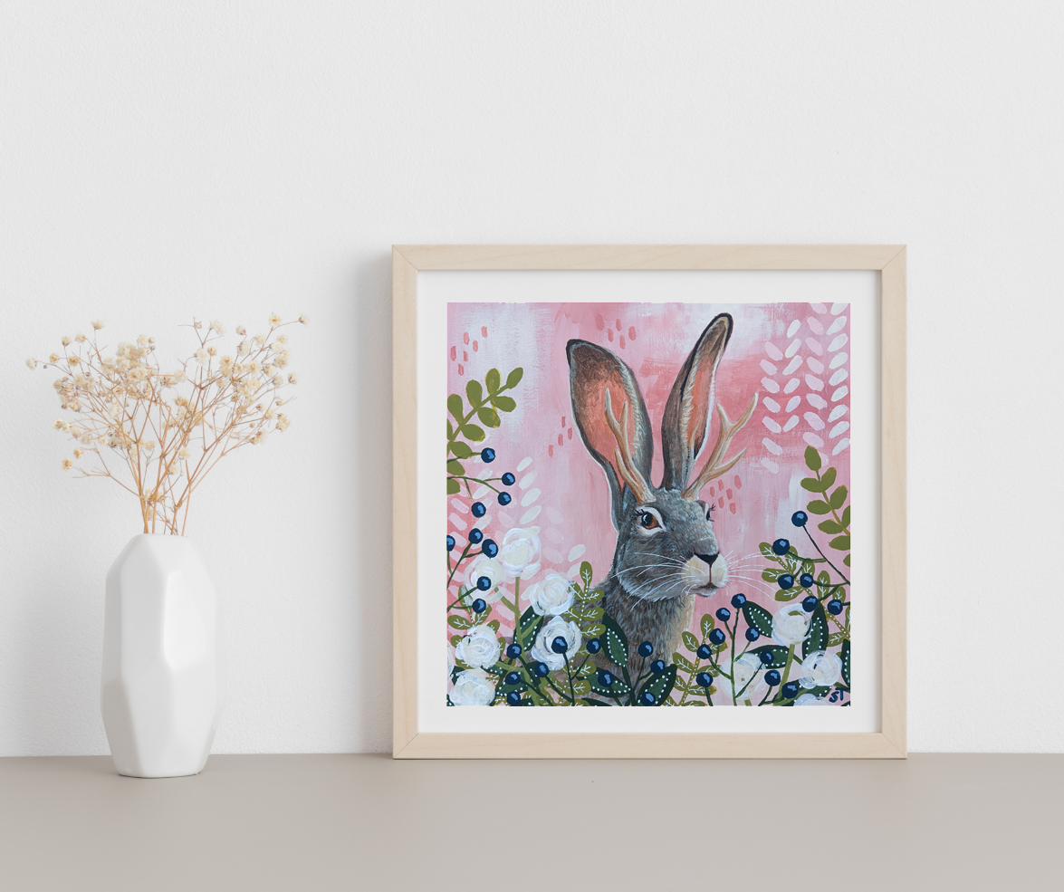 Framed cryptid art print of a mythical jackalope with branching antlers rising from a floral foreground, set against a soft pink folkloric backdrop, displayed on a minimalist shelf.