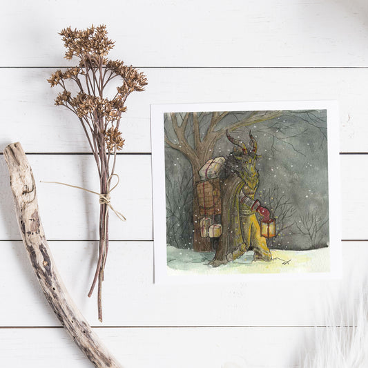 Note card featuring and krampus  in a winter scene, next to dried flowers and a branch on a white wooden surface.