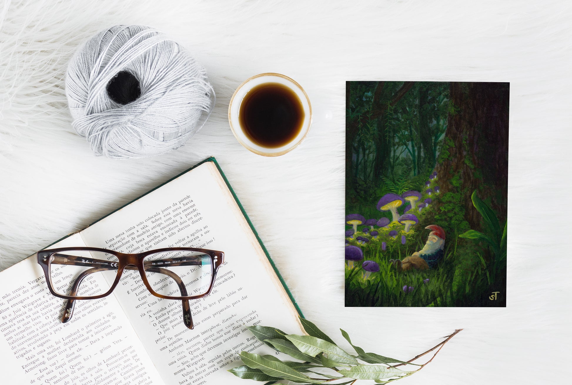 Sleeping gnome resting among glowing mushrooms in a mossy forest at night, painted in rich greens and purples, shown as a greeting card styled with a book and coffee.