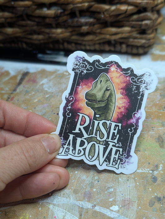 Sticker with a dinosaur design and 'Rise Above' text held by a hand on a textured surface.