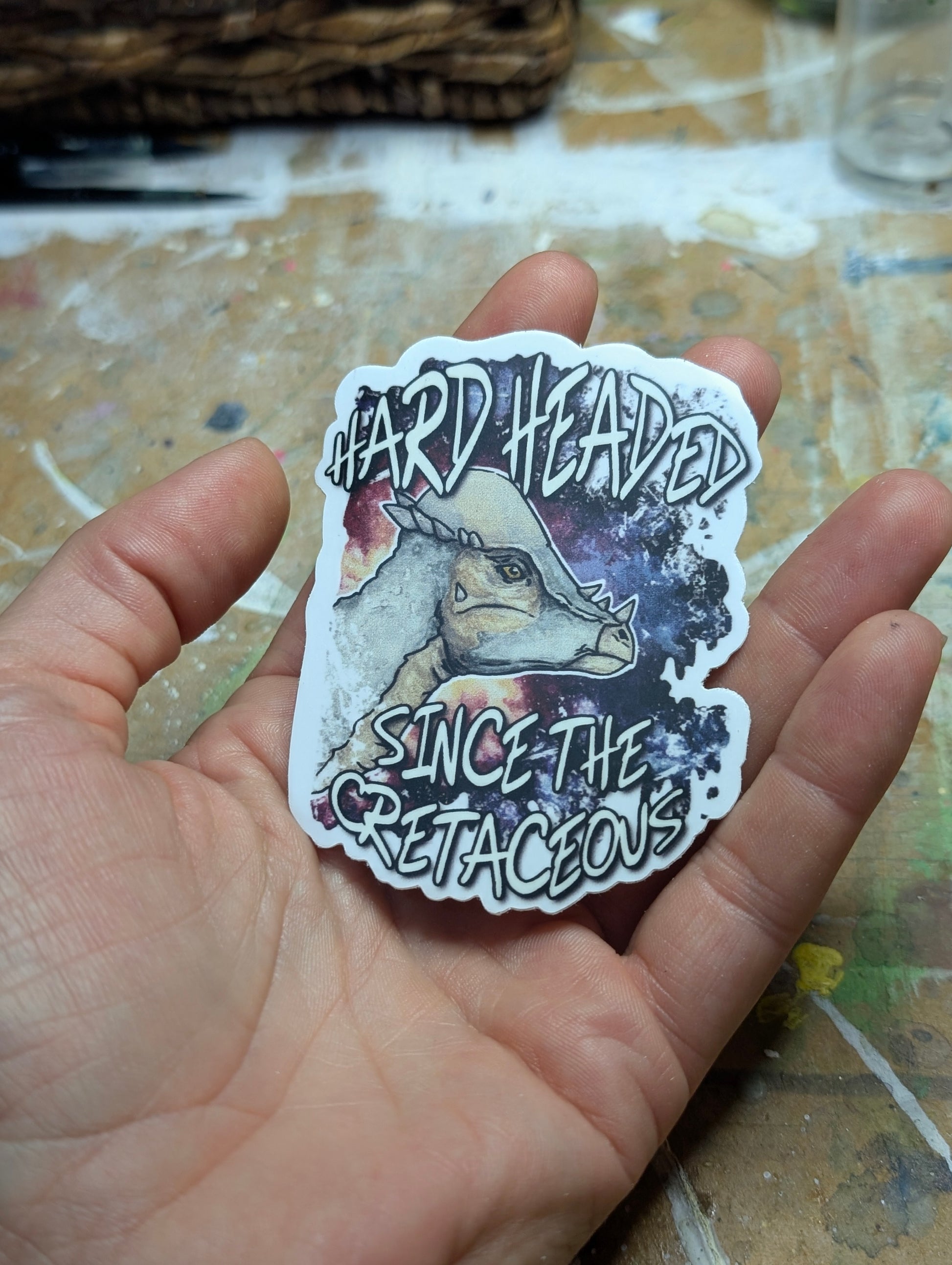 Sticker with a pachycephalosaurus design held in a hand on a textured surface.