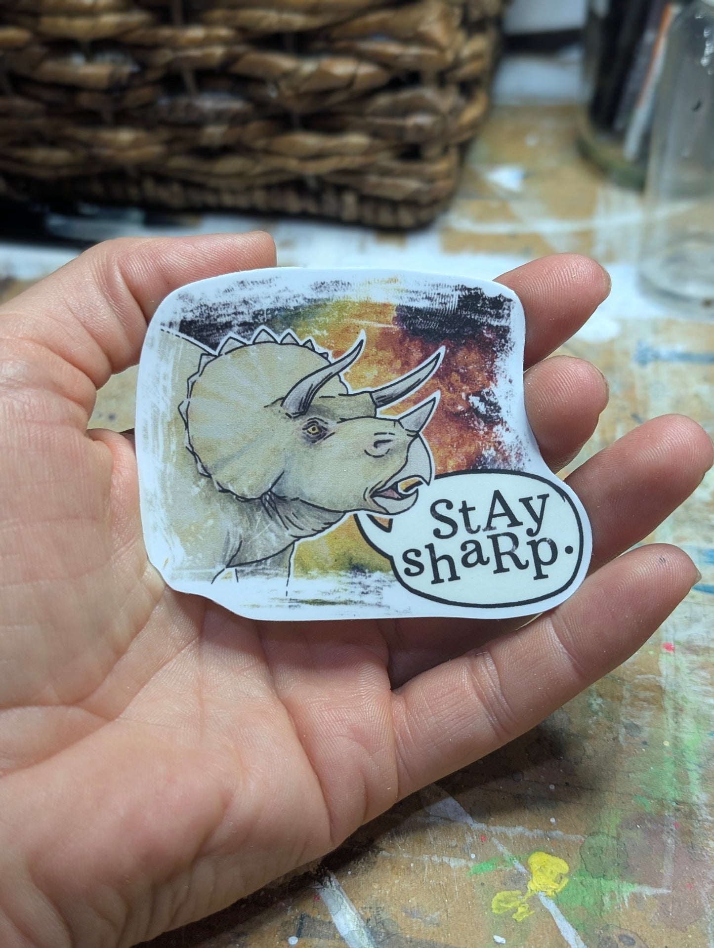 Hand holding a sticker with a triceratops illustration and 'Stay sharp' text.
