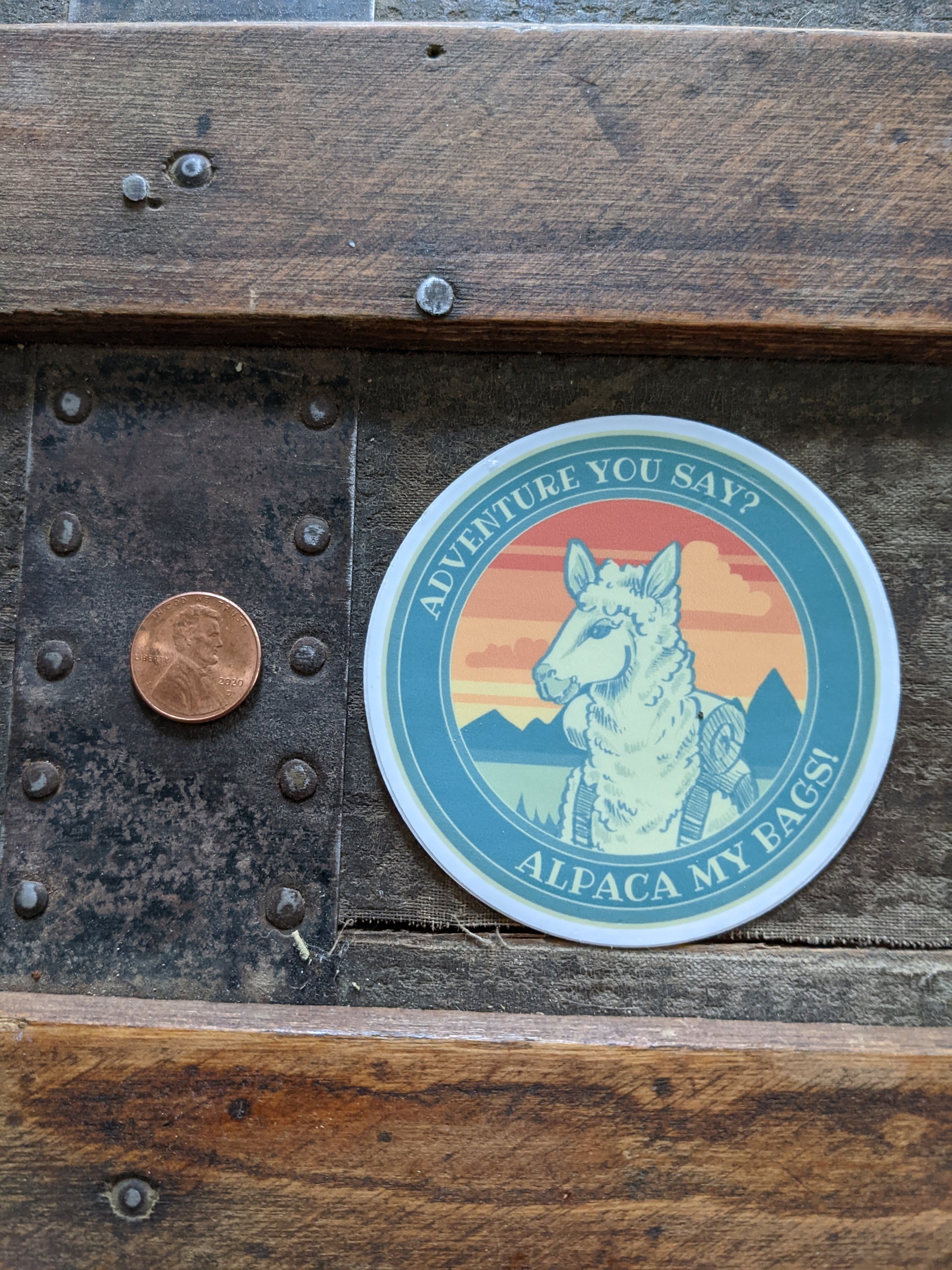 Round alpaca vinyl sticker shown next to a penny for size reference, featuring mountain scenery and “Alpaca my bags” text.