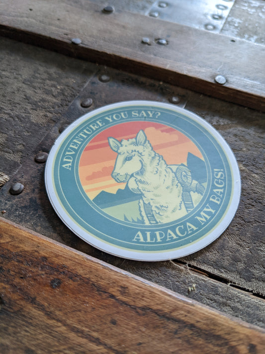 Round vinyl sticker featuring an illustrated alpaca wearing hiking gear with mountains and a sunset background and the text “Adventure you say? Alpaca my bags!”