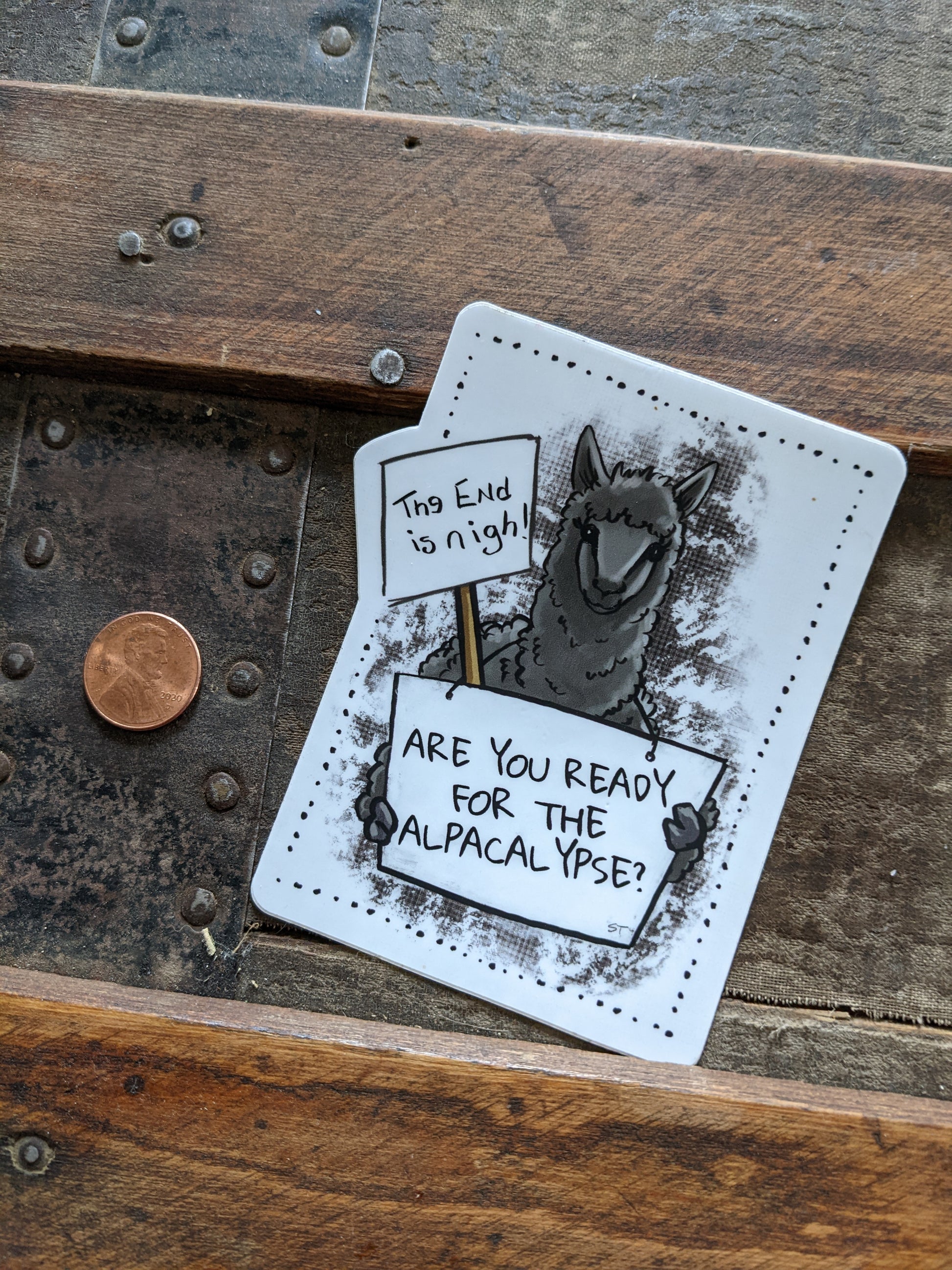 Alpaca “alpacalypse” vinyl sticker shown next to a penny for size reference, featuring protest-style signs with humorous text.