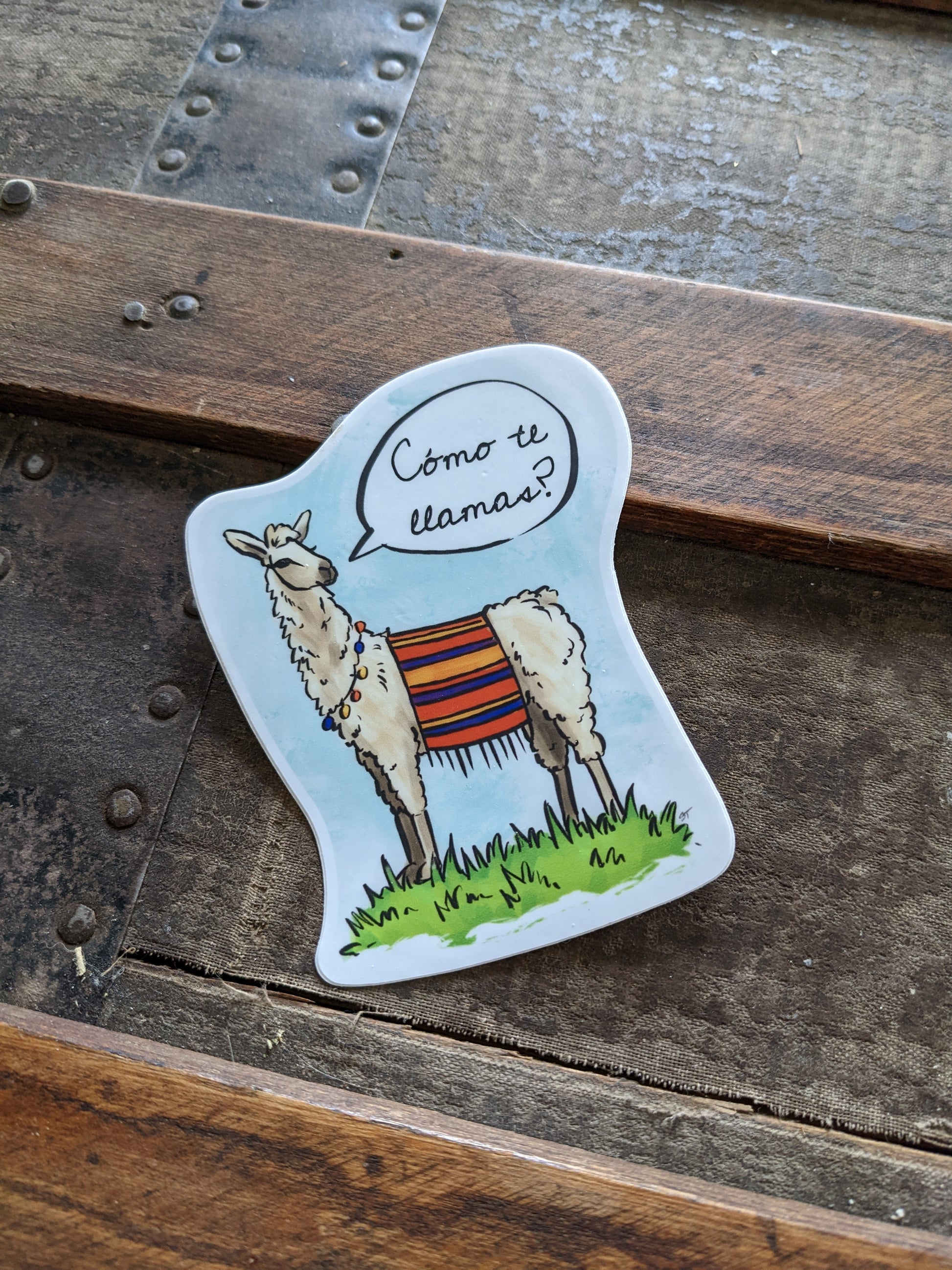 Vinyl sticker featuring an illustrated llama wearing a colorful blanket and necklace, with a speech bubble reading “¿Cómo te llamas?”