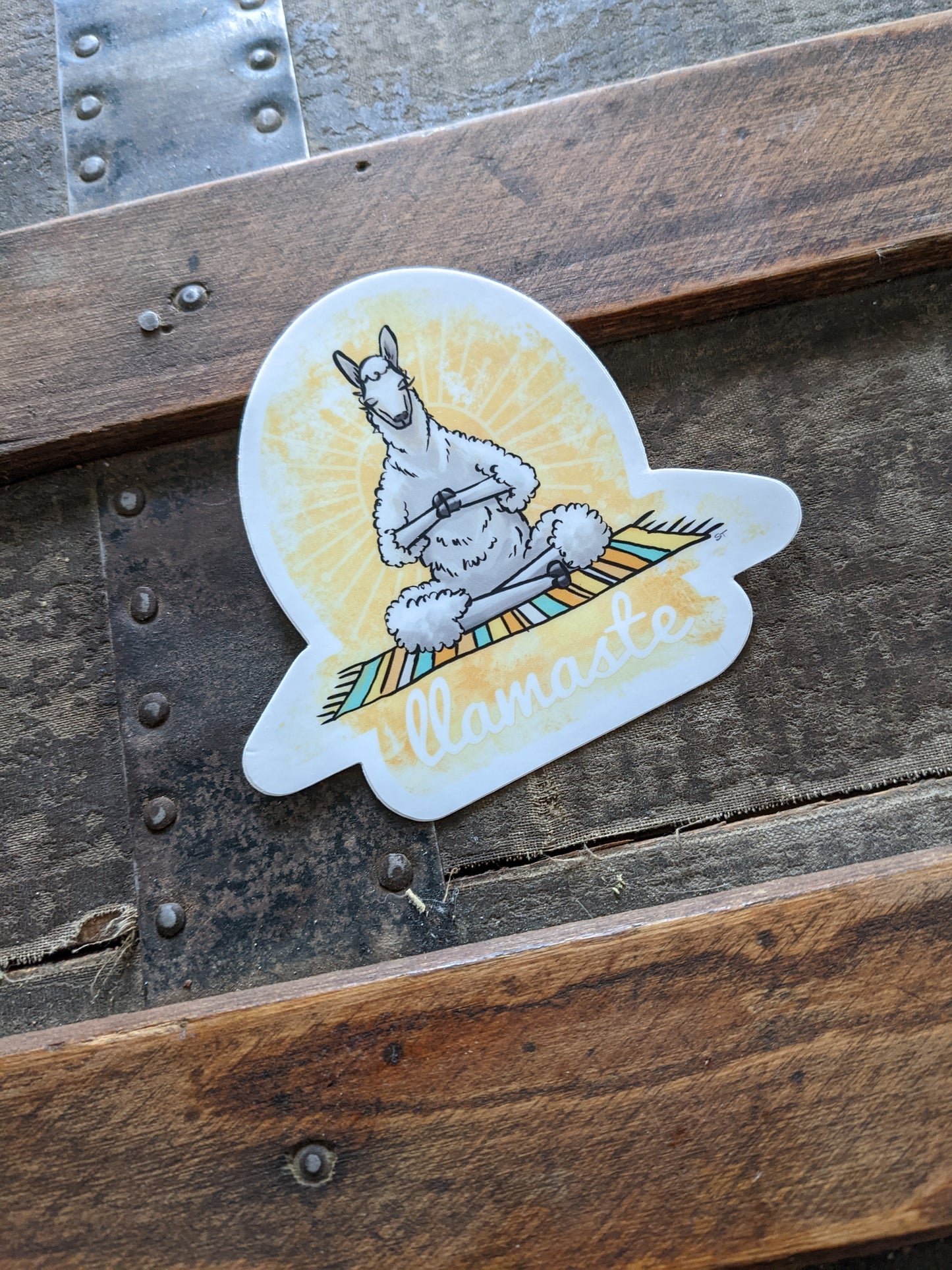 “Llamaste” vinyl sticker featuring a meditating llama on a striped mat, photographed on an old steamer trunk.