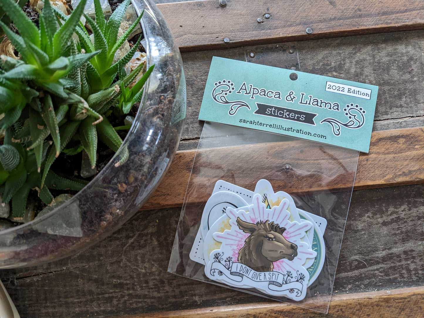 A set of alpaca and llama themed stickers with puns, displayed on a wooden surface next to a glass bowl with a plant.