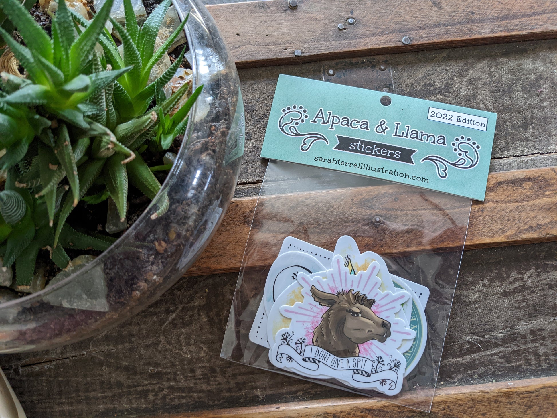 A set of alpaca and llama themed stickers with puns, displayed on a wooden surface next to a glass bowl with a plant.
