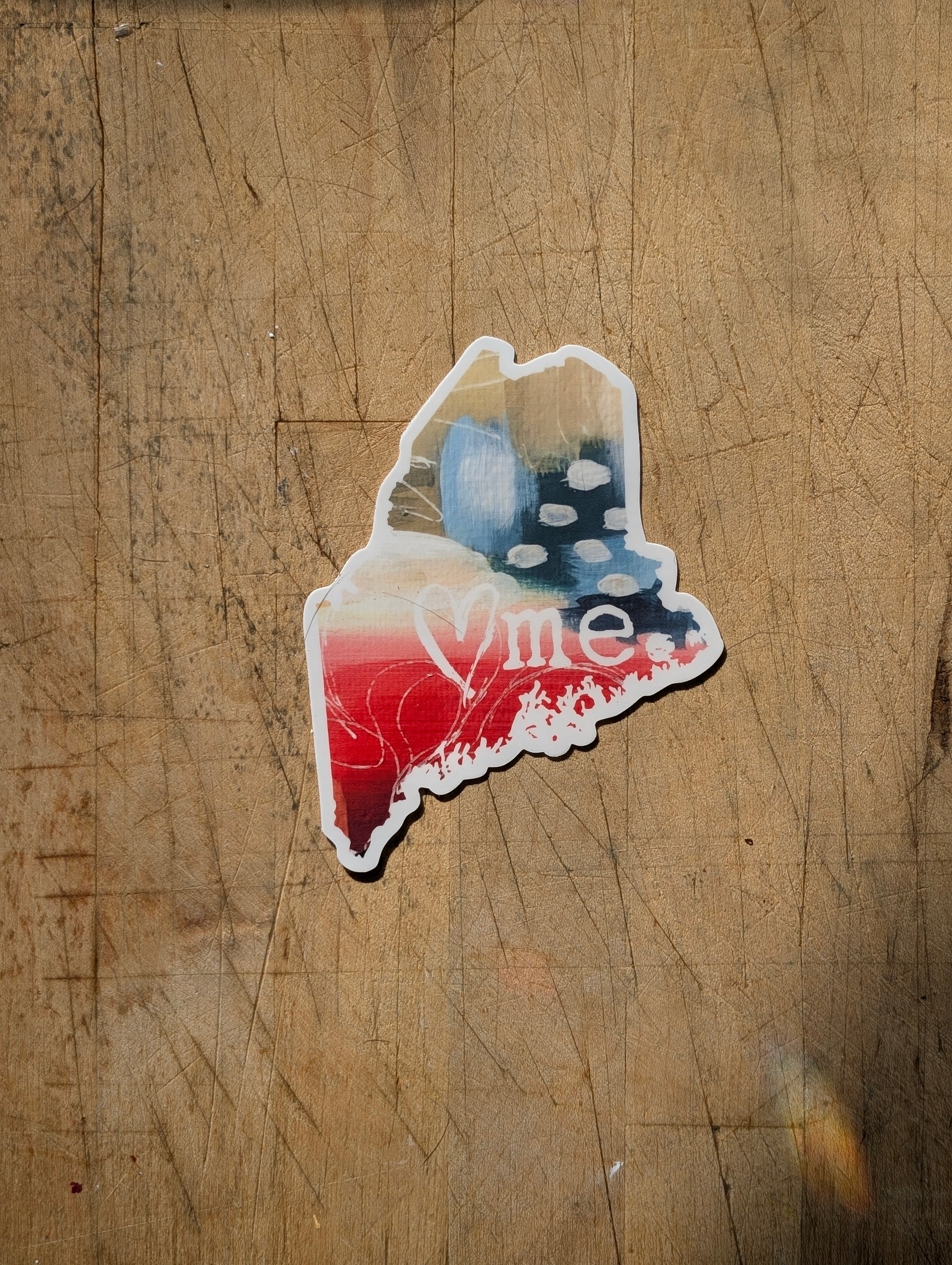 Abstract Maine-shaped “heart ME” vinyl sticker with red and teal painted textures, shown in a flat lay on wood.