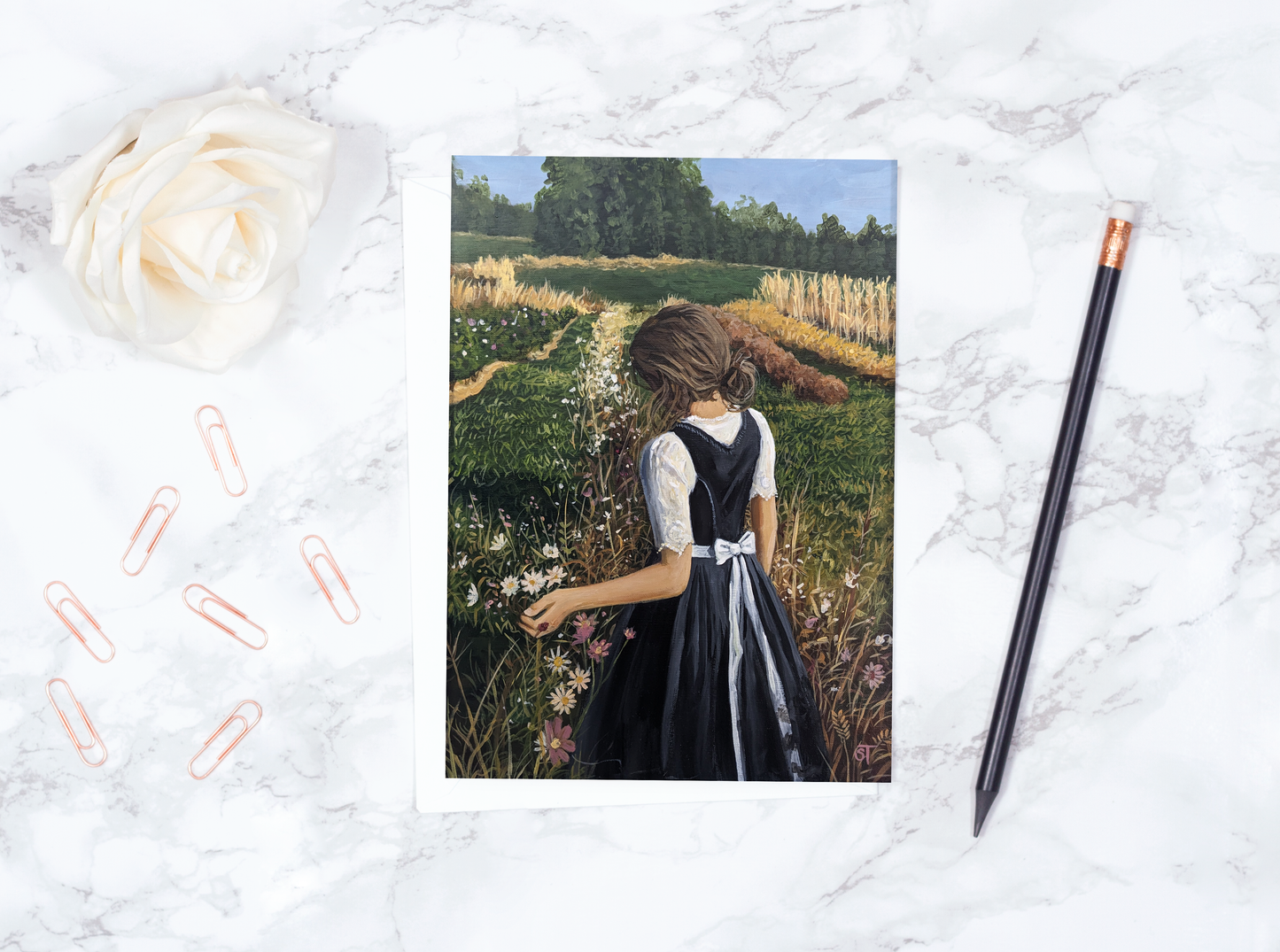 A greeting card featuring an acrylic painting of a girl walking through a field of flowers, with a brown kraft envelope and a pencil on a marble surface.