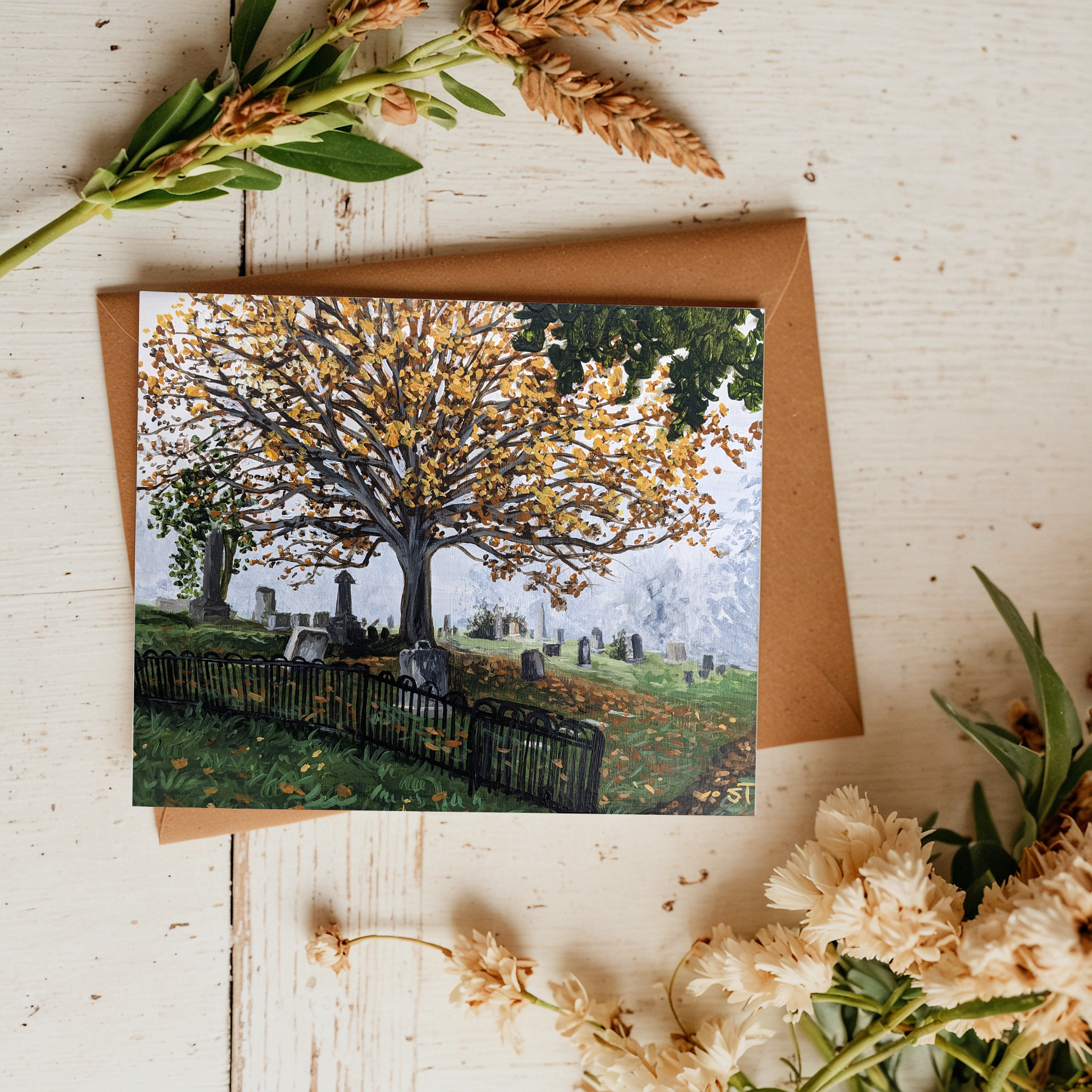 A greeting card featuring an autumnal landscape with a tree and gravestones, presented on a wooden surface with dried flowers nearby.