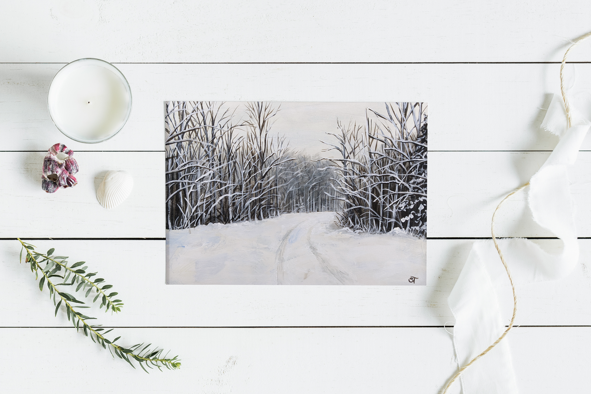 A greeting card featuring a winter landscape with snow-covered trees and a rural road, displayed on a white wooden surface with a candle and decorative foliage in the background.