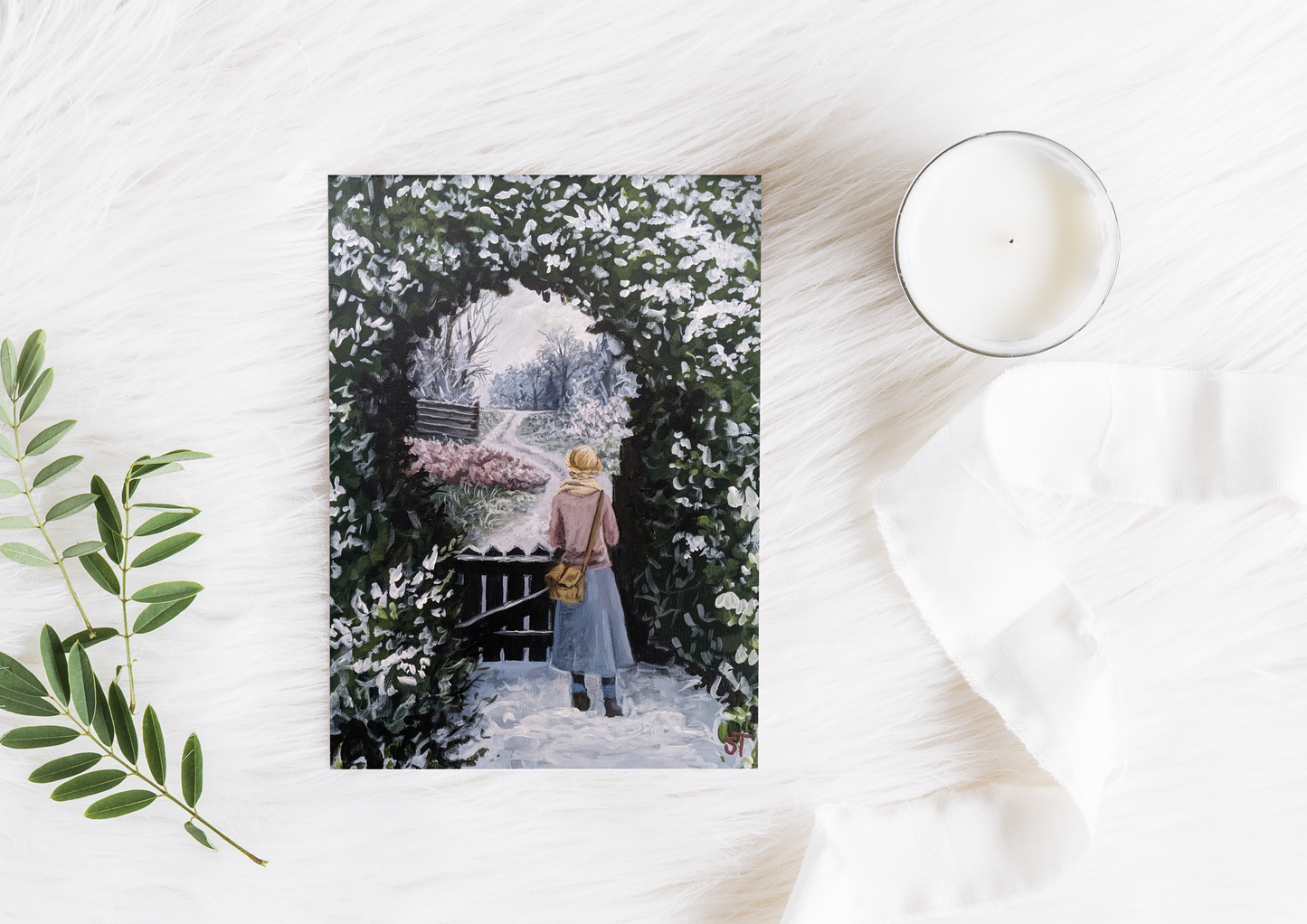 A greeting card featuring an acrylic painting of a girl standing by a garden gate, with frosted trees and a cup of tea in the foreground, placed on a white surface with a candle and decorative leaves.
