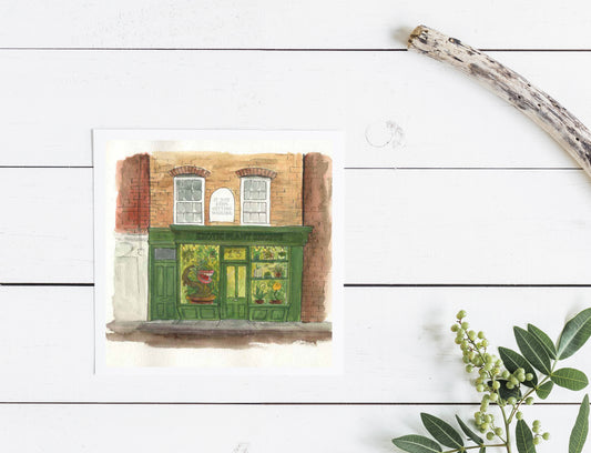 Small greeting card featuring a painting of an exotic plant shop with a green facade, displayed on a wooden surface with a branch and greenery.