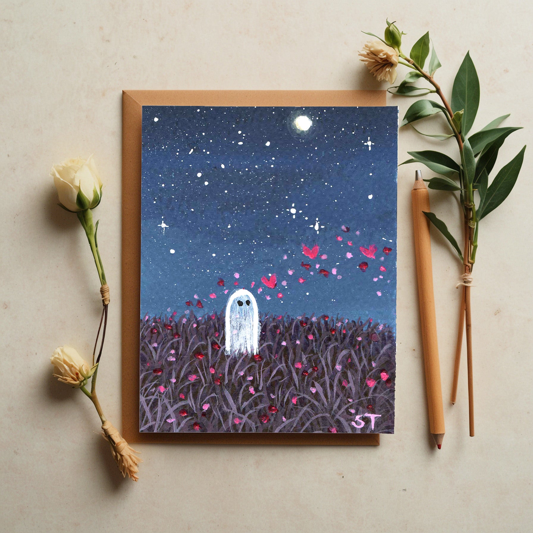 Greeting card with ghost and starry night design, surrounded by flowers and pencils on a beige surface.