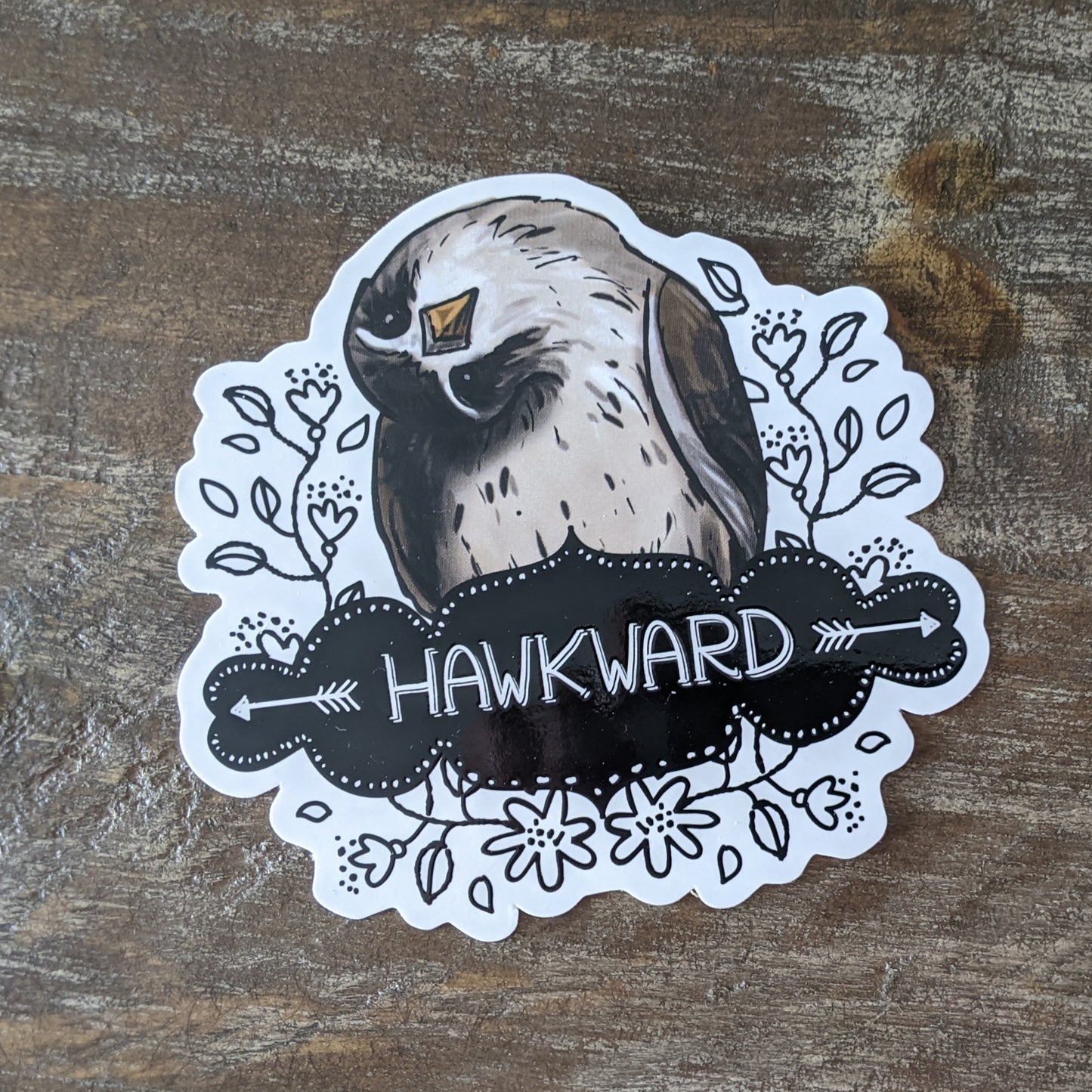 A sticker with an illustration of a bird and the word 'HAWKWARD' written on it, placed on a wooden surface.