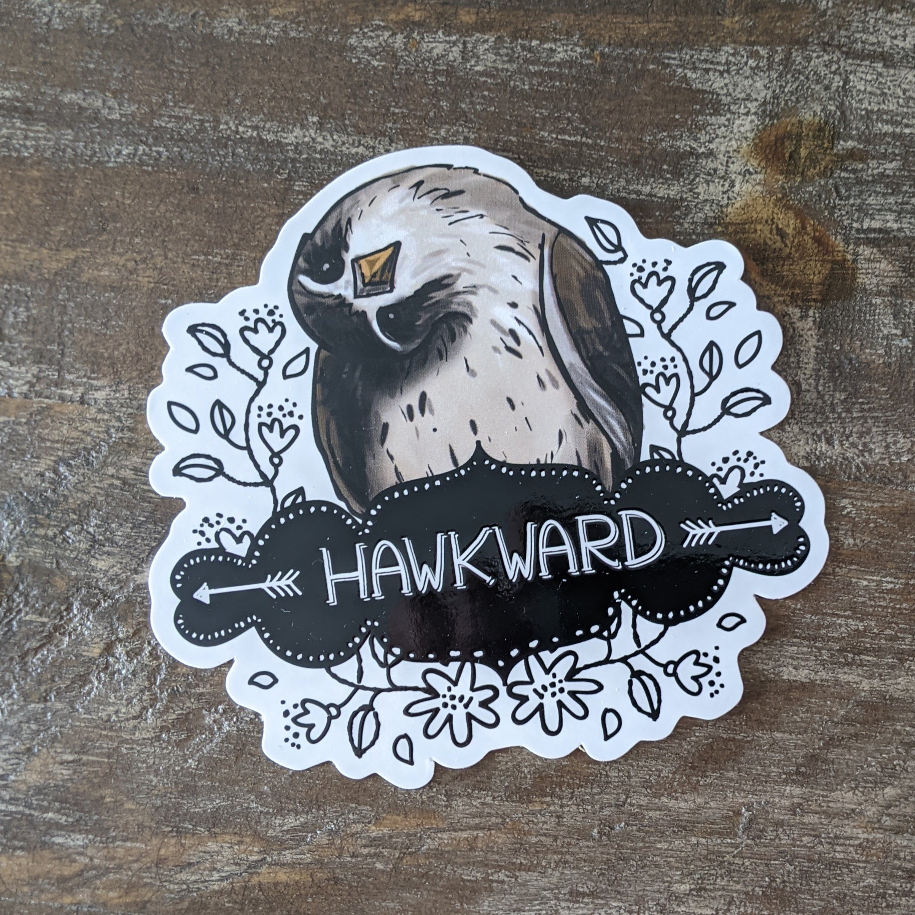 A sticker with an illustration of a bird and the word 'HAWKWARD' written on it, placed on a wooden surface.