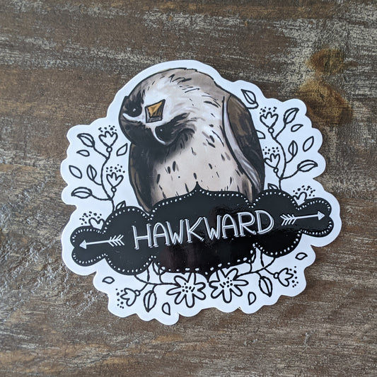 A sticker with an illustration of a bird and the word 'HAWKWARD' written on it, placed on a wooden surface.