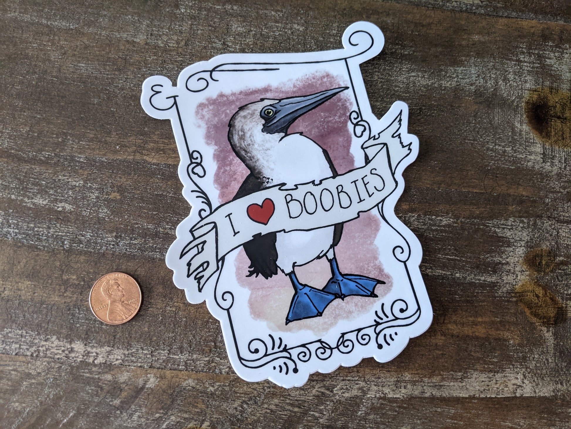 Blue-Footed Booby vinyl sticker with a banner reading “I ♥ boobies,” shown next to a penny for size reference on a wooden surface.