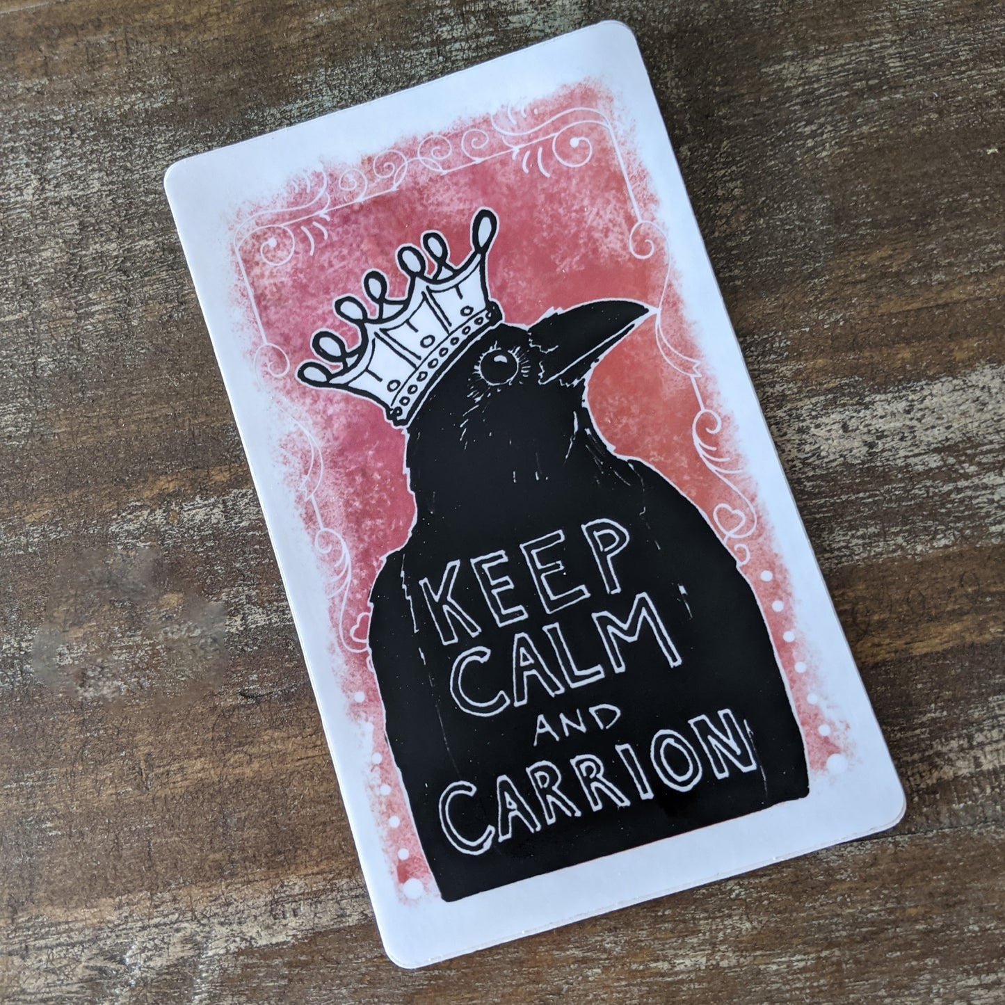 A vinyl sticker with a illustration of a raven wearing a crown, with the text 'KEEP CALM AND CARRION' on a wooden surface.