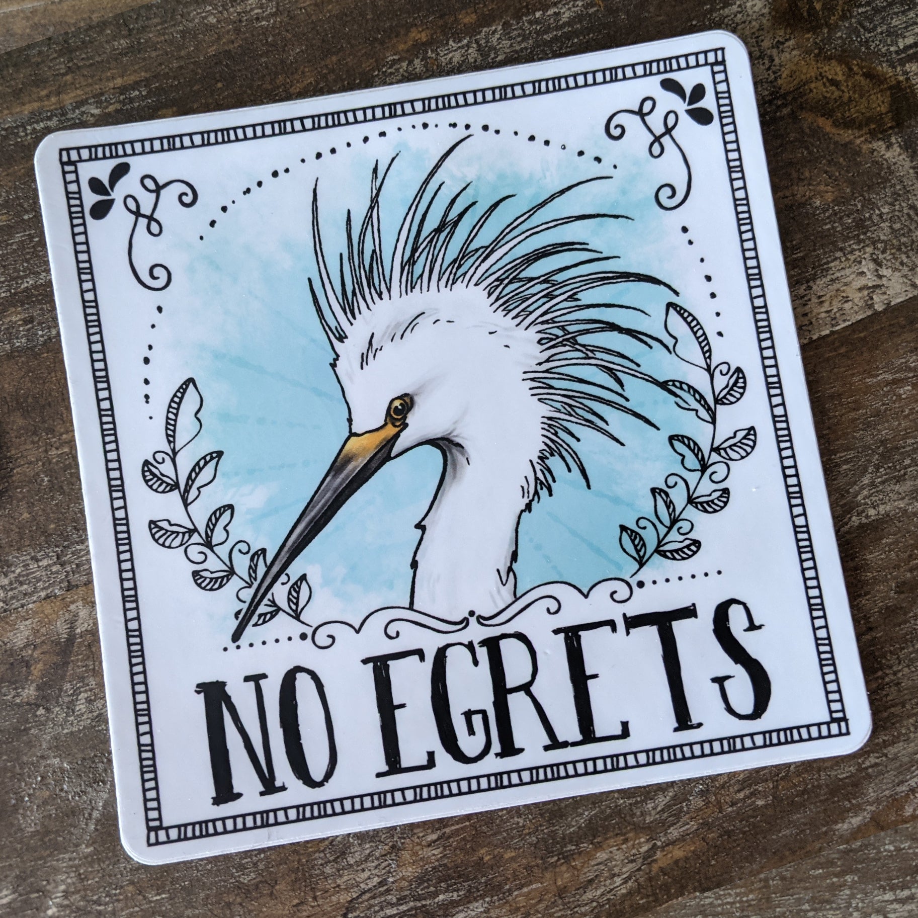 A vinyl decal featuring an illustration of an egret with the text 'NO EGrets' playfully arranged lain on a wooden surface.