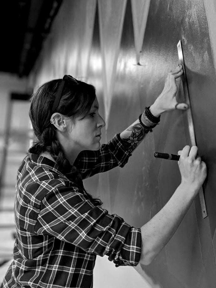 Black and white photo of the artist Sarah Terrell measuring and marking a large wall mural with a ruler and paint marker while wearing a plaid shirt.