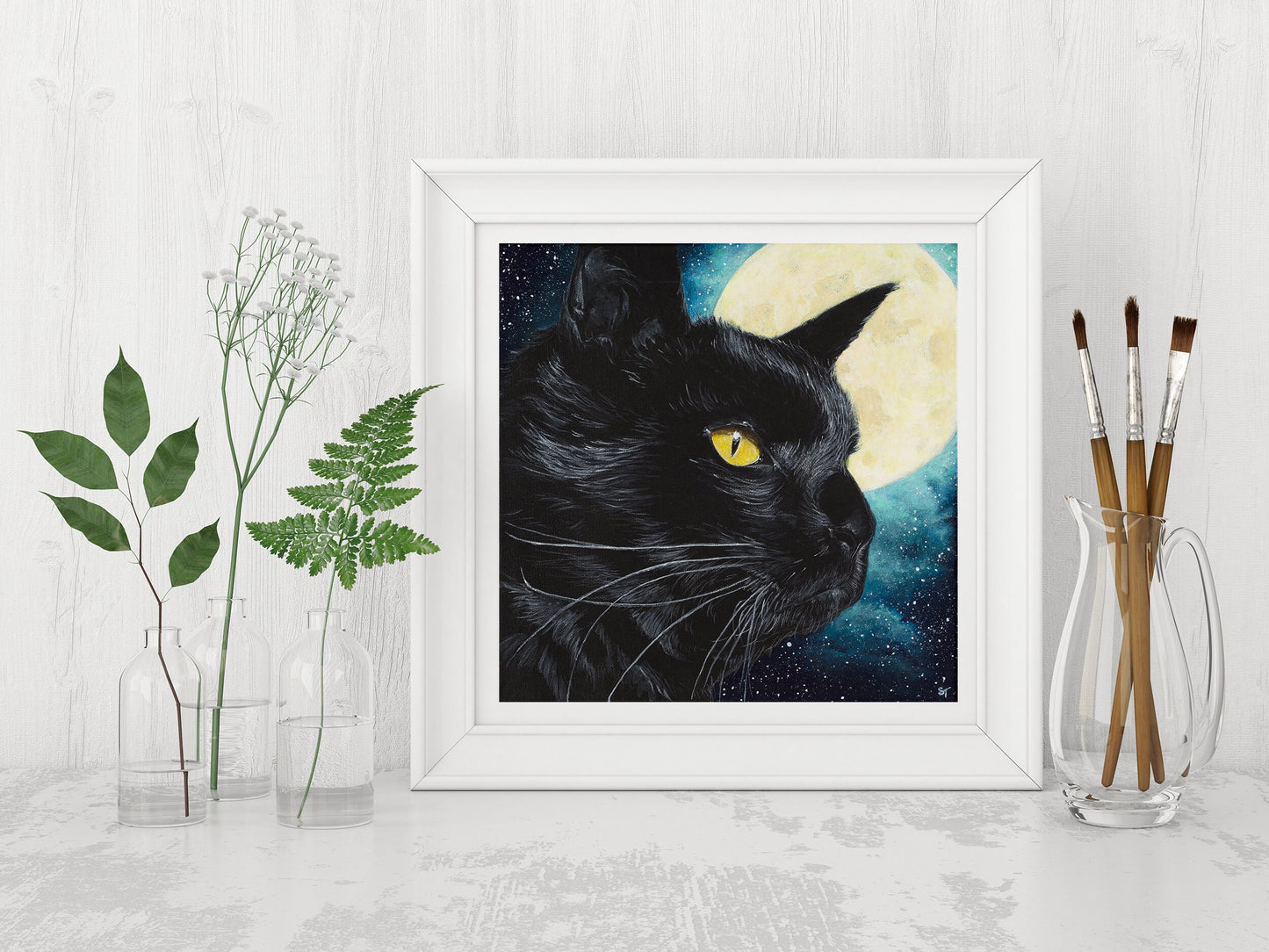 A framed art print of a black cat with a moon and stars in the background, displayed on a white wall with a plant and jar of paint brushes to the side.