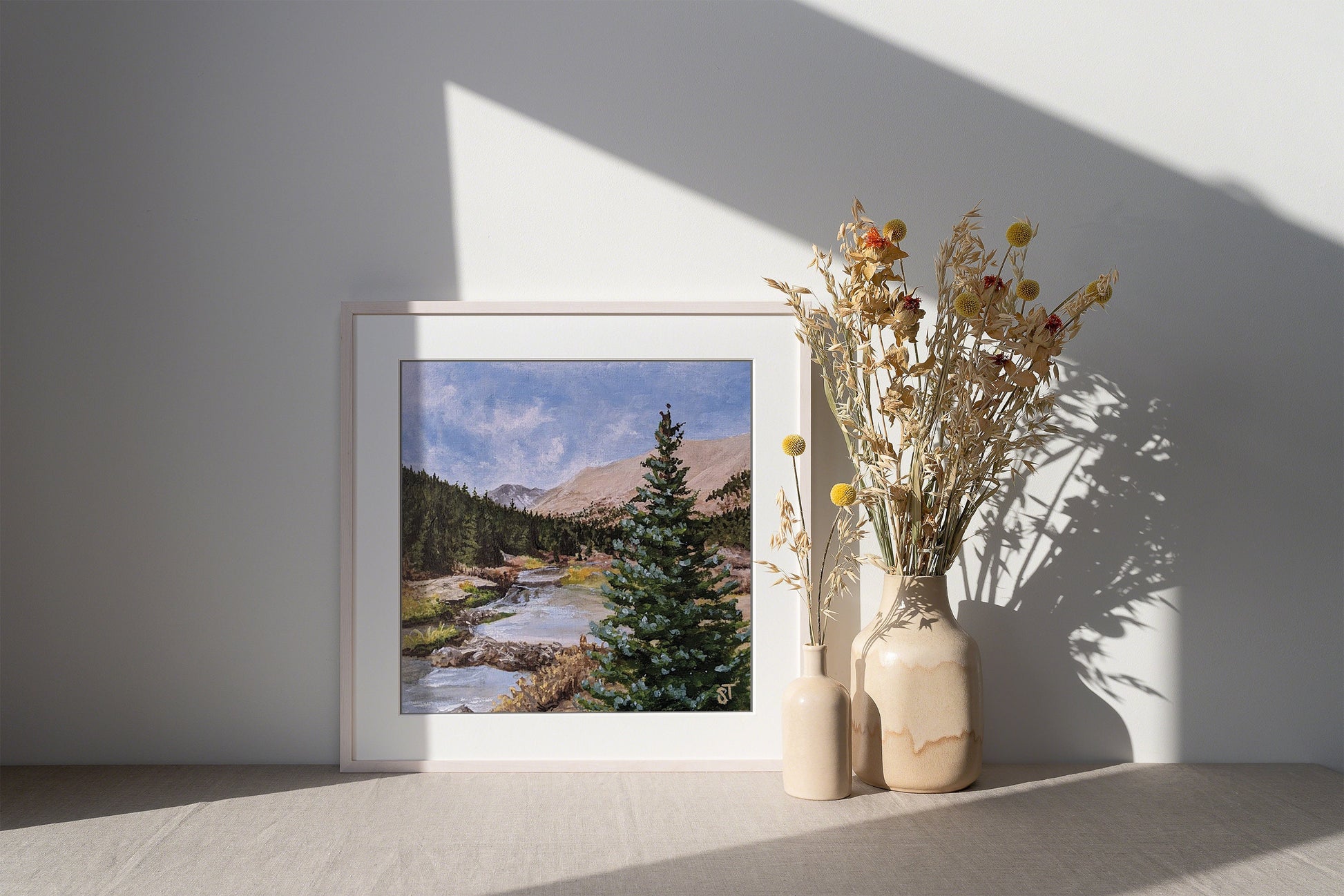 A framed art print featuring a landscape with a river, trees, and mountains, placed on a wooden surface next to a vase with dried flowers.