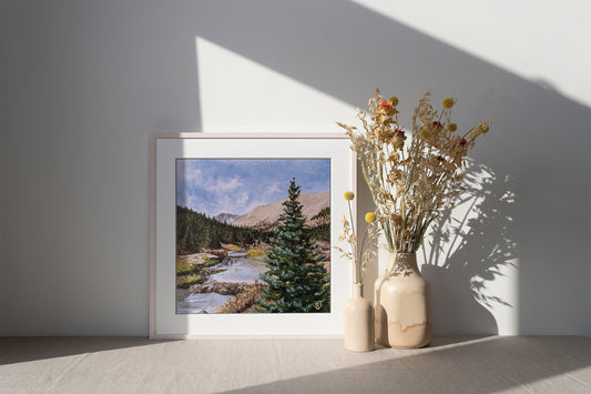 A framed art print featuring a landscape with a river, trees, and mountains, placed on a wooden surface next to a vase with dried flowers.