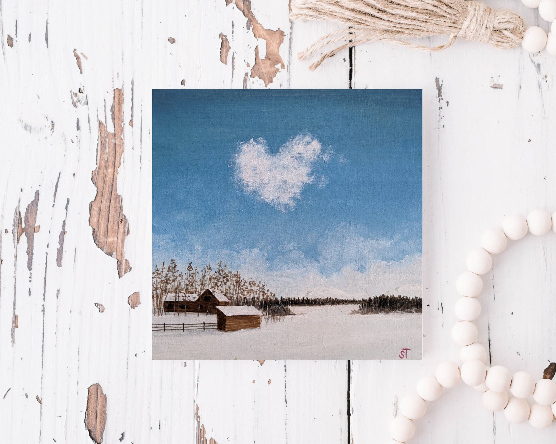 Flat lay of a winter landscape greeting card showing a snow-covered field, rustic barn, and a heart-shaped cloud in a blue sky, styled with soft neutral décor.